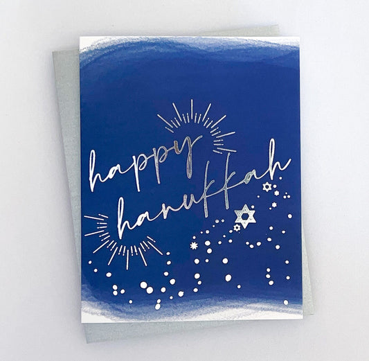 A blue card with 'Happy Hanukkah' text in silver foil, along with star and light designs.