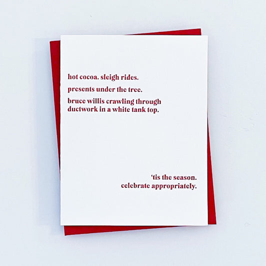 A holiday letterpress card with a red envelope, featuring the text 'Hot cocoa. Sleigh rides. Presents under the tree. bruce willis crawling through ductwork in a white tank top.' 'tis the season . celebrate appropriately.'