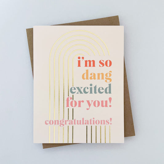 A greeting card with gold foil accent in a rainbow shape, colorful text that reads 'I'm so dang excited for you!' 'congratulations!' and paired with a brown kraft envelope.