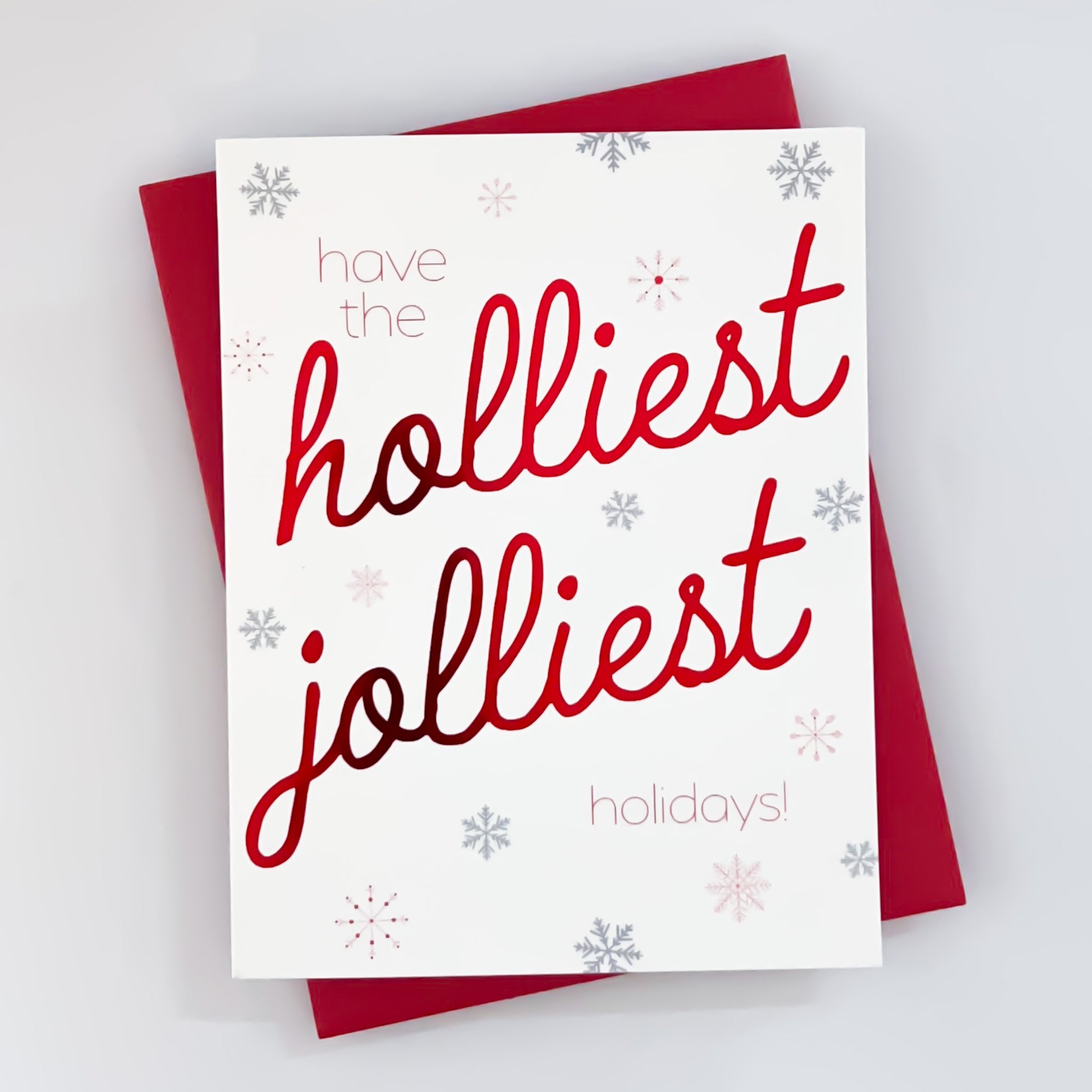 A holiday card with white background with snowflakes with red foil text that reads 'have the holliest jolliest holidays'