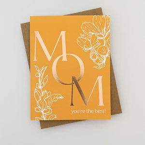 A burnt orange greeting card with floral patterns and 'Mom' written in a large, decorative font, accompanied by 'You're the best!' and some copper foil details.