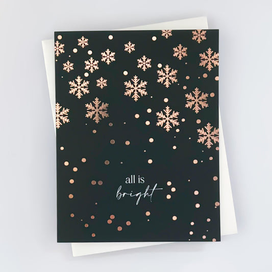 A holiday card with a dark green background and rose gold foil snowflake patterns, featuring an envelope and the text 'All is bright' on the card.