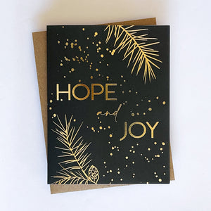 A holiday card with a dark green background and gold foil accents, featuring the text 'Hope and Joy' along with pine tree sprigs and dot design.