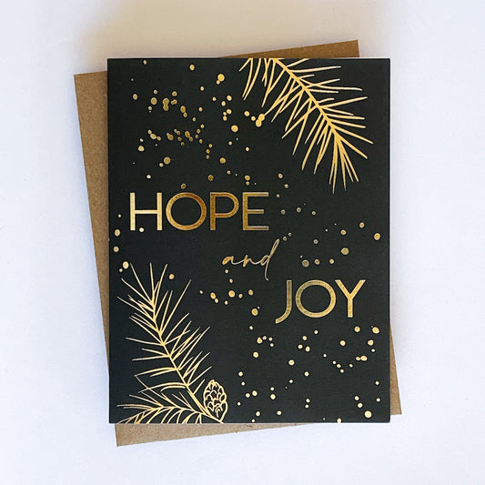 A holiday card with a dark green background and gold foil accents, featuring the text 'Hope and Joy' along with pine tree sprigs and dot design.