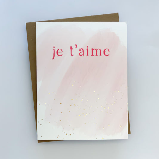 A greeting card with a watercolor illustration of pink and gold foil dots on it, with the text 'Je t'aime' printed in red.