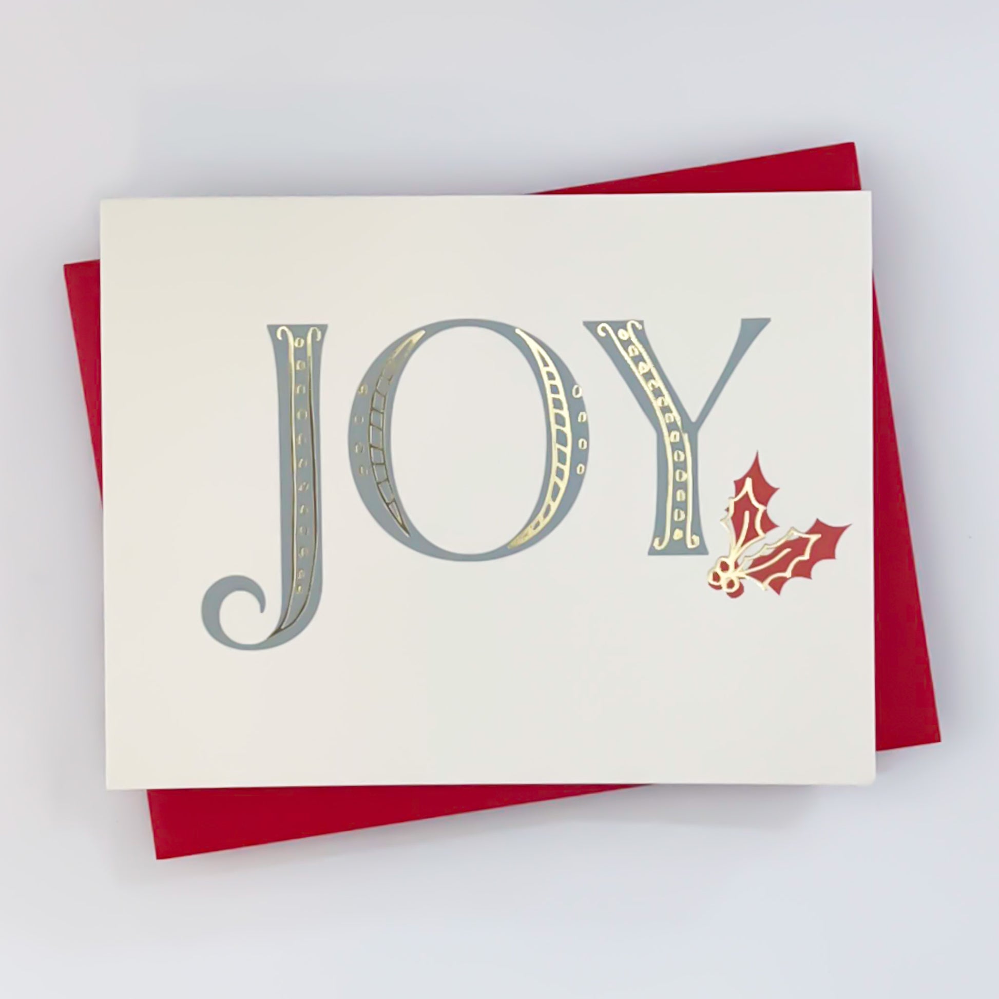 A holiday card with white background and text that writes 'JOY' across the front with red holly design and gold foil accents 