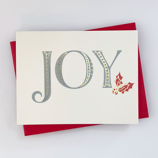 A holiday card with white background and text that writes 'JOY' across the front with red holly design and gold foil accents 