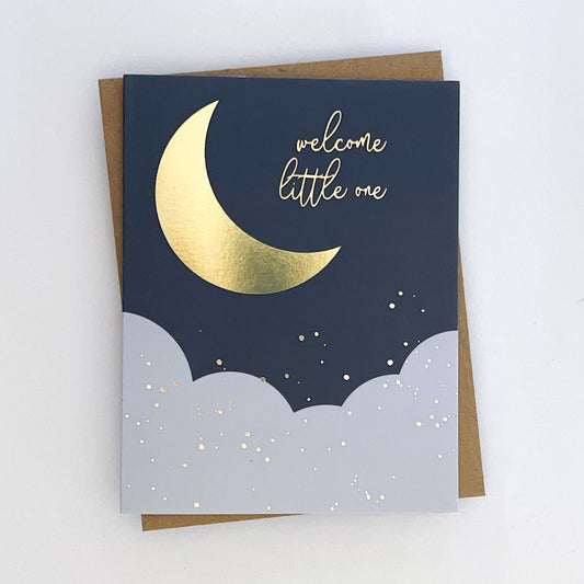 A gold foil card with a moon and stars design on a dark background, with the text 'Welcome little one' printed in gold. The card is presented with a kraft envelope.