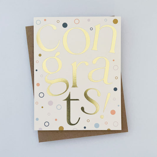 A greeting card with a cream color background, featuring gold foil lettering that reads 'CONGRATS!' with colorful accents and a kraft envelope.