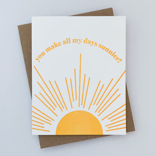 A greeting card with a sun and sunbeams design in yellow, with the text 'You make all my days sunnier!', accompanied by a Kraft envelope.