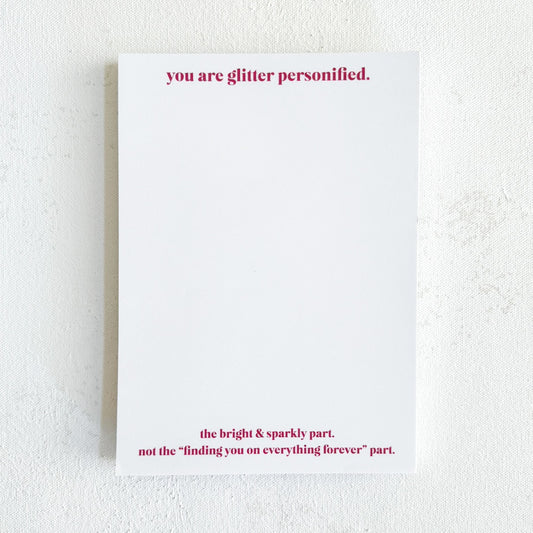 A white notepad with bright fuschia text at the top that reads 'you are glitter personified' and text at the bottom that reads 'the bright & sparkly part. not the "finding you on everything forever" part'