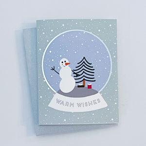 A greeting card with a winter scene that includes a snowman, a Christmas tree, and the text 'Warm Wishes' on the front, with silver foil details on the card.
