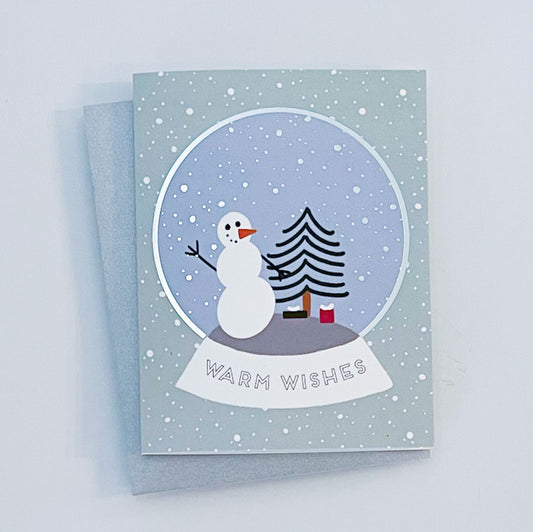 A greeting card with a winter scene that includes a snowman, a Christmas tree, and the text 'Warm Wishes' on the front, with silver foil details on the card.