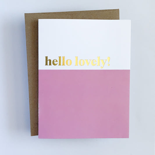 A greeting card with a white background and gold foil text that reads 'Hello Lovely!' The card has a pink colorblock design and is paired with a brown kraft envelope.