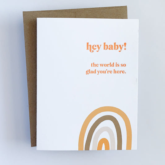A Letterpress Baby Shower Card with text that reads 'hey baby! the world is so glad you're here.' written in orange complimented by and orange and neutral tone rainbow. 