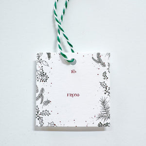 A white gift tag with to and from text in red surrounded be black holly and winter greenery paired with a green and white string.