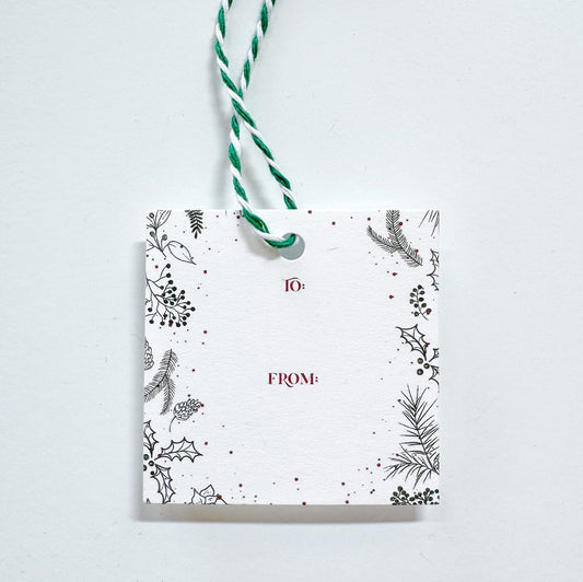 A white gift tag with to and from text in red surrounded be black holly and winter greenery paired with a green and white string.