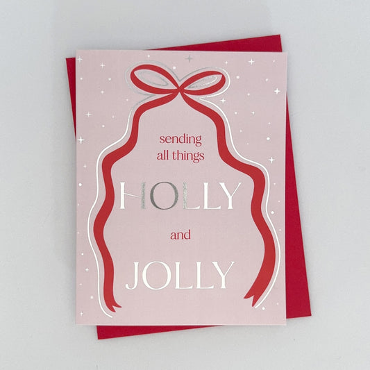 A holiday card with a pink background, featuring a hand-drawn red ribbon bow and silver foil accents, with the text 'SENDING ALL THINGS HOLY AND JOLLY'. 