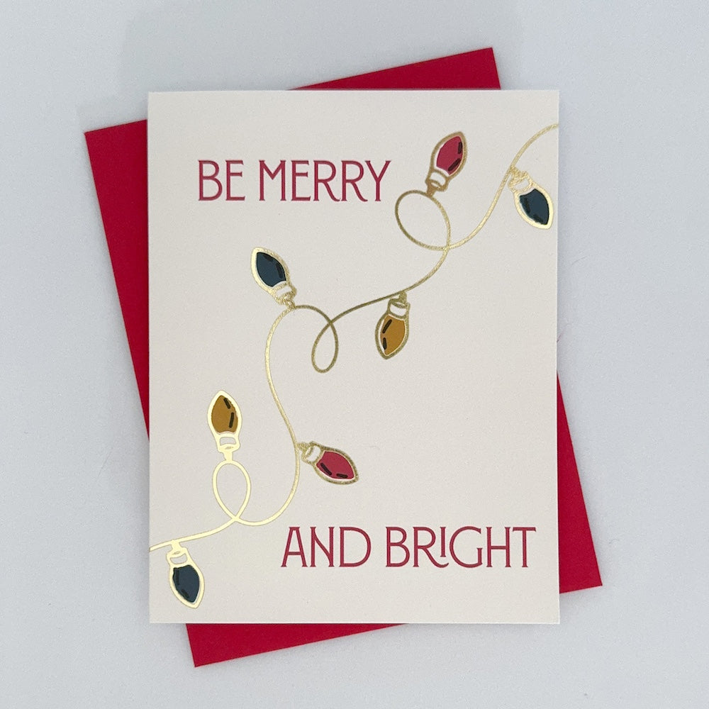 A holiday card with hand-drawn light illustrations and gold foil accents, featuring an message that reads 'Be Merry And bright'. The card has a red envelope and is presented against a white background.