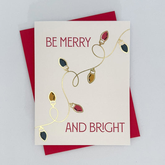 A holiday card with hand-drawn light illustrations and gold foil accents, featuring an message that reads 'Be Merry And bright'. The card has a red envelope and is presented against a white background.