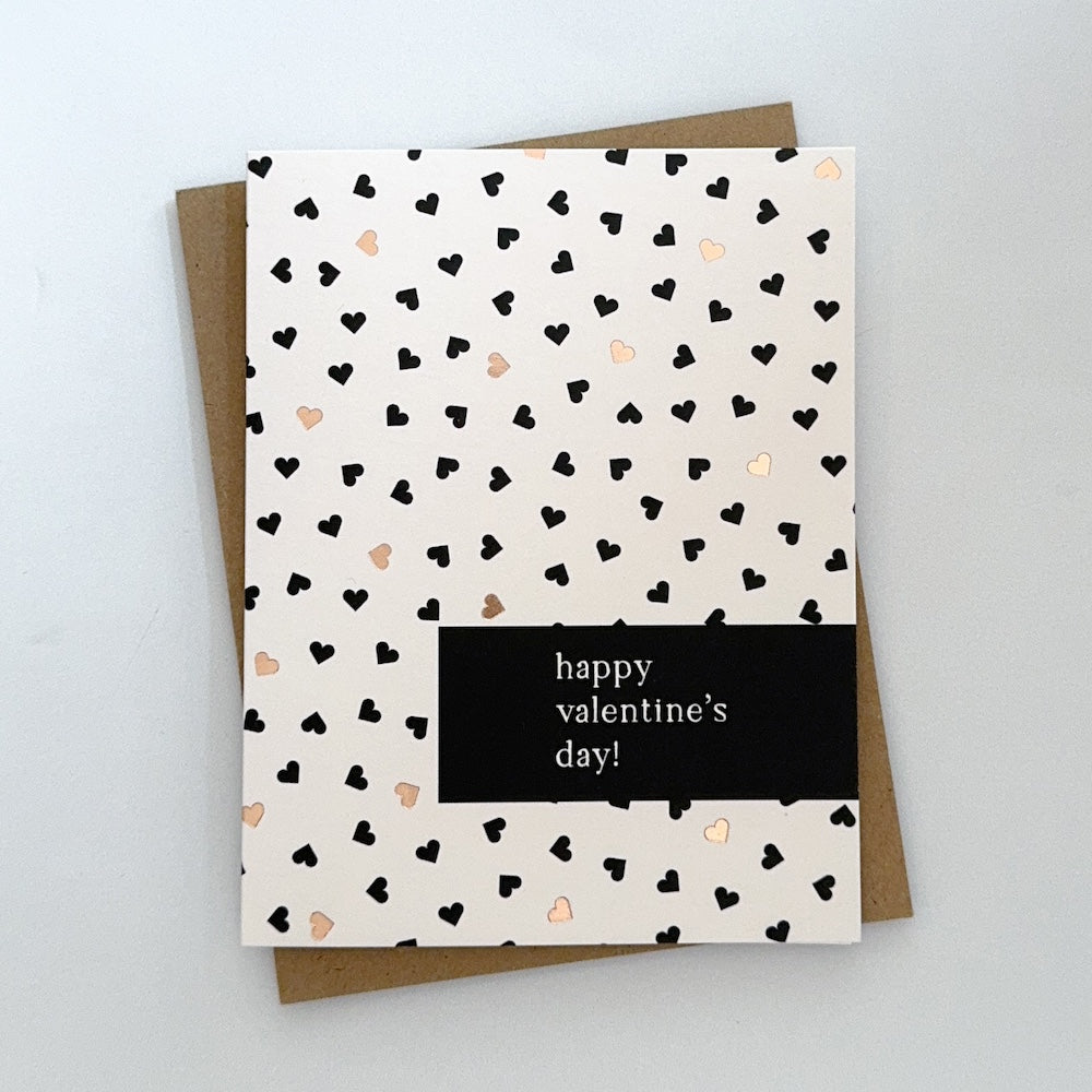 A Valentines Day card with a light pink background with tiny black and gold foil hearts. The text reads 'Happy Valentines Day'.