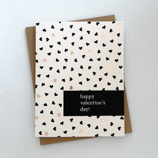 A Valentines Day card with a light pink background with tiny black and gold foil hearts. The text reads 'Happy Valentines Day'.