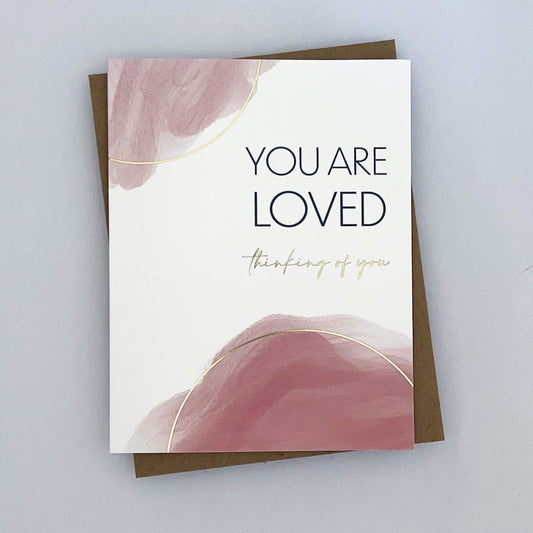 A greeting card with a watercolor print in shades of pink, gold foil accents, and a blank interior for a personal message. The card reads 'You are loved thinking of you'.
